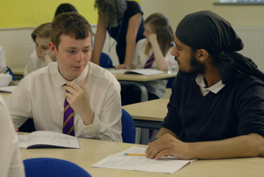 White male teen and BAME male teen talk at school desk looking concerned